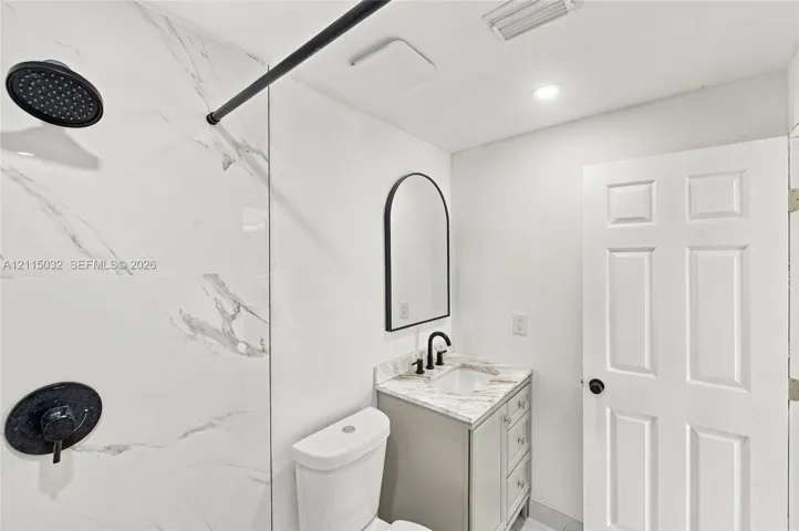 Bathroom featuring a shower with large-format patterned wall tile and matte black fixtures, a neutral vanity with a stone-finish countertop, and an arched wall mirror