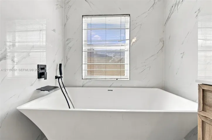 Freestanding rectangular soaking tub featuring large-format porcelain wall tile with a veined pattern, matte black wall-mounted faucet and handheld shower sprayer, and a built-in recessed shelf