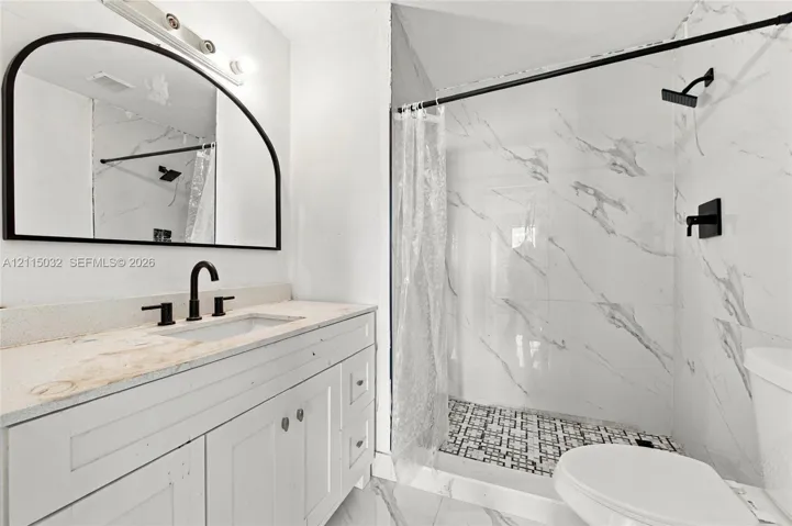 Bathroom featuring a white vanity cabinet, light-toned countertop with integrated sink, arched wall-mounted mirror, and walk-in shower with large-format patterned wall tile
