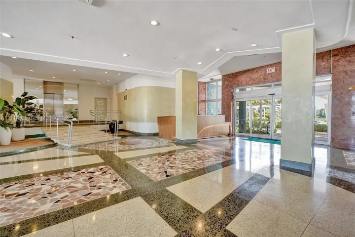 Grand lobby featuring multi-textured stone flooring, high ceilings with recessed lighting, structural support columns, a built-in reception desk, and glass-paned sliding entry doors