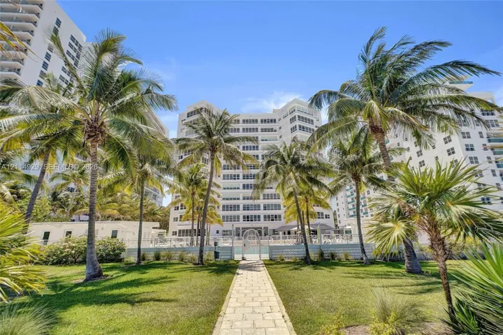 High-rise exterior with white stucco facade, stone-paver walkway, manicured lawn, and mature palm trees