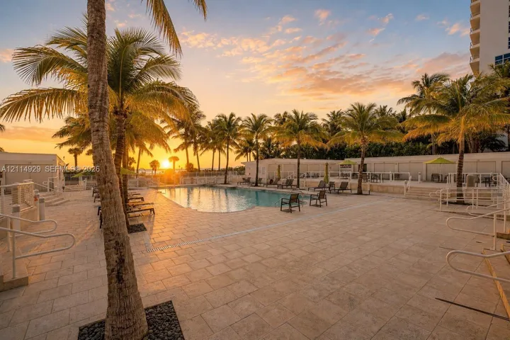 Swimming pool area featuring expansive stone tile decking, mature palm trees, perimeter metal fencing, and integrated pool entry stairs, set against a sunset backdrop