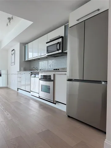 Kitchen featuring high-gloss cabinetry, stainless steel appliances, light-toned wood-finish flooring, and a subway tile backsplash