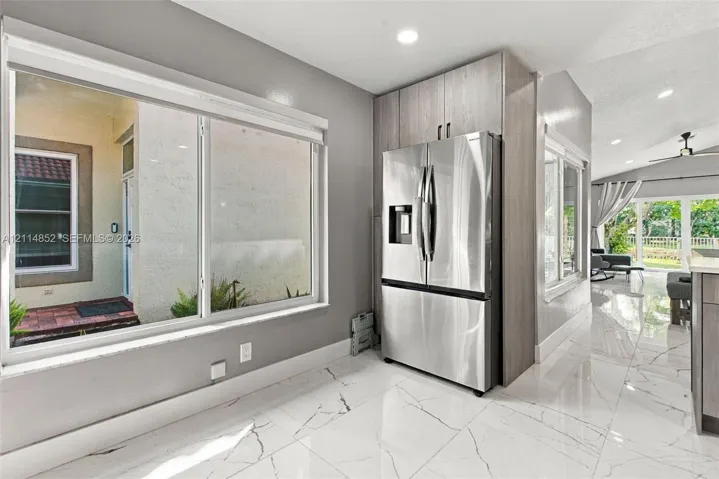 Large picture window, stainless steel French-door refrigerator, floor-to-ceiling wood-finish cabinetry, and polished stone-finish tile flooring with veining