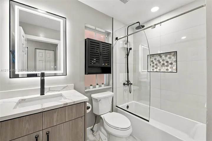 Bathroom featuring a wood-finish vanity with a stone-top integrated sink, matte black faucet, LED-backlit mirror, and a tub-shower combination with a glass panel and hexagon tile recessed niche