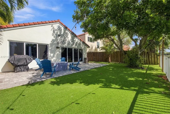 Backyard featuring a paved stone patio, synthetic lawn, and mature trees, enclosed by perimeter fencing