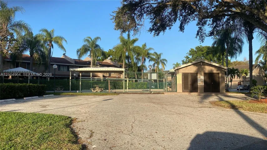 Community pool area featuring perimeter fencing, a covered canopy structure, a stand-alone utility building, and mature palm trees