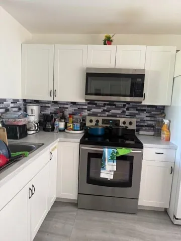 Kitchen featuring white shaker-style cabinetry, stainless steel appliances, multi-tonal linear tile backsplash, solid-surface countertops, and large-format tile flooring