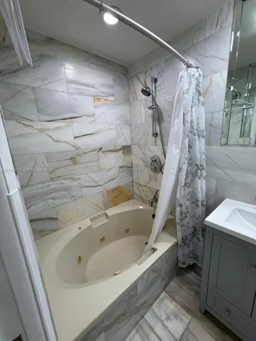Jetted soaking tub with wall-mounted chrome hand shower, surround of large-format stone-patterned tile, and grey built-in vanity with an integrated sink