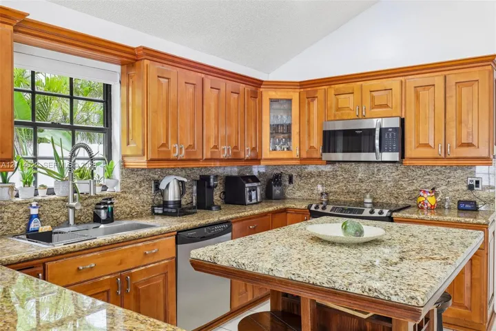 Kitchen featuring warm-toned wood cabinetry, expansive stone countertops, a multi-tier island, a stainless steel sink with an arched faucet, and built-in stainless steel appliances