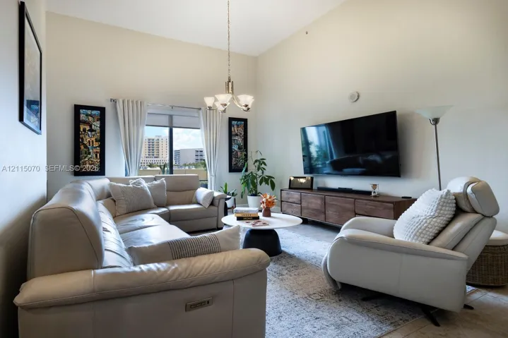 High vaulted ceiling with neutral paint palette, large window with natural light, multi-arm chandelier, and stone-finish flooring