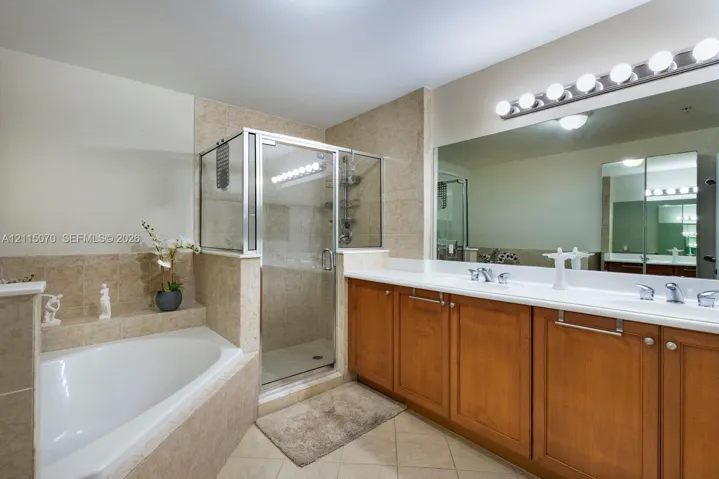 Bathroom featuring a dual-sink vanity with wood cabinetry, white countertops, glass-enclosed shower, built-in bathtub with tiled surround, and expansive wall-mounted mirror