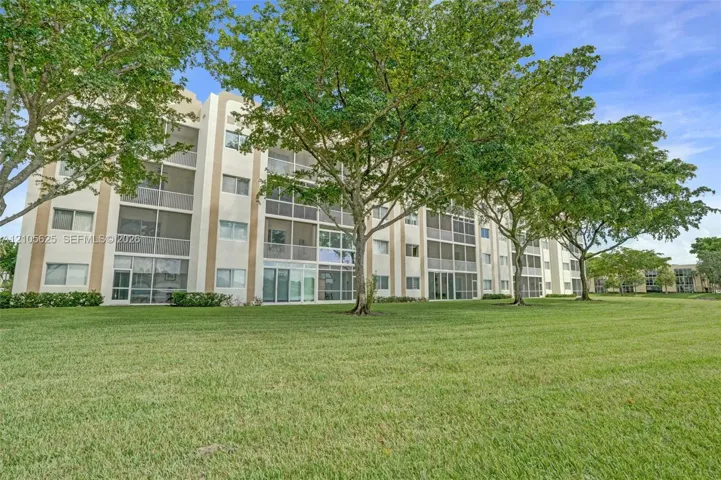 Multi-story building exterior featuring light-toned stucco facade, screened balconies, and extensive surrounding lawn with mature trees