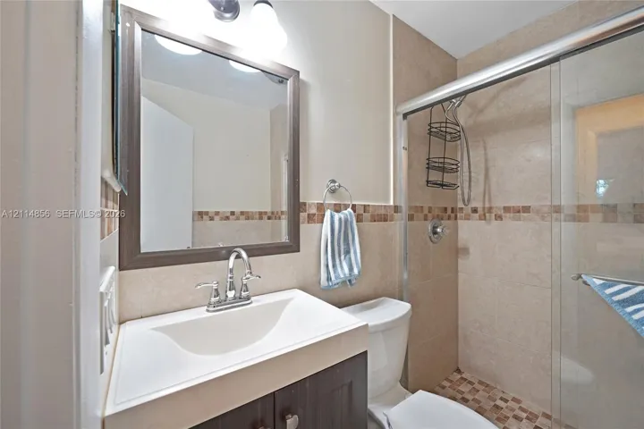 Bathroom featuring a dark wood-finish vanity with an integrated white sink, framed wall mirror, tiled shower with a sliding glass door, and decorative mosaic tile accents