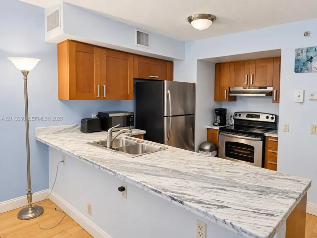 Kitchen featuring wood-finish cabinetry, stone-patterned countertops, a stainless steel double-basin sink, and integrated stainless steel appliances