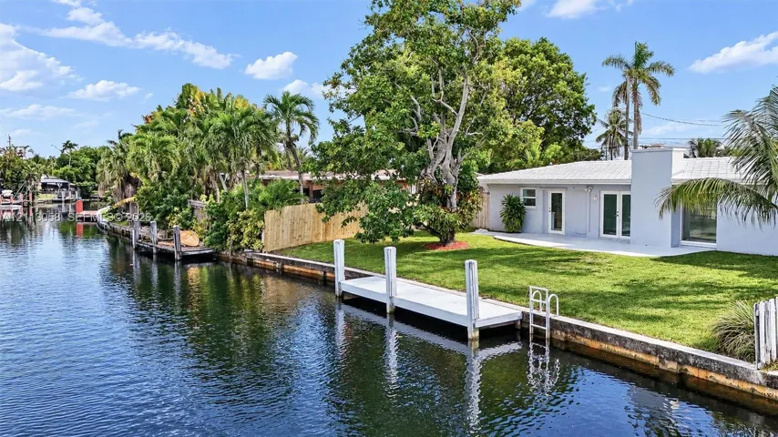 Waterfront property featuring a private dock, concrete seawall, expansive lawn, mature palm trees, and a ground-level patio