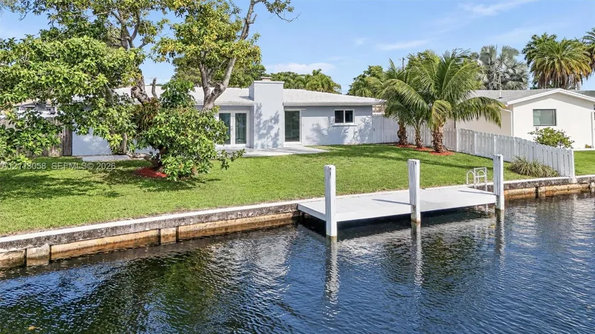 Waterfront residence featuring a private boat dock, concrete seawall, expansive lawn, and white vinyl perimeter fencing