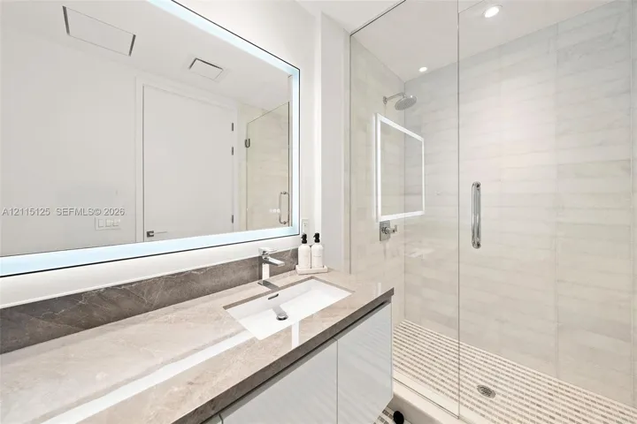 Floating vanity with stone countertop, rectangular undermount sink, integrated backlit mirror, and glass-enclosed shower with tiled flooring