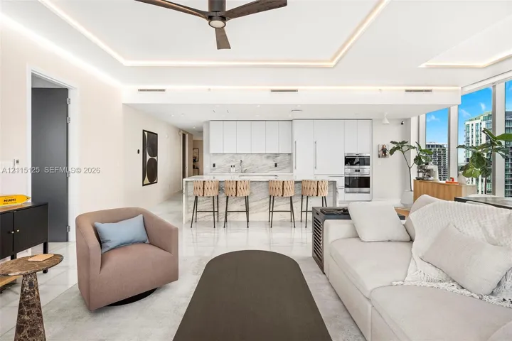 Open-concept interior featuring recessed tray ceilings with cove lighting, expansive floor-to-ceiling windows, stone-surround kitchen island, and custom flat-panel cabinetry with integrated appliances
