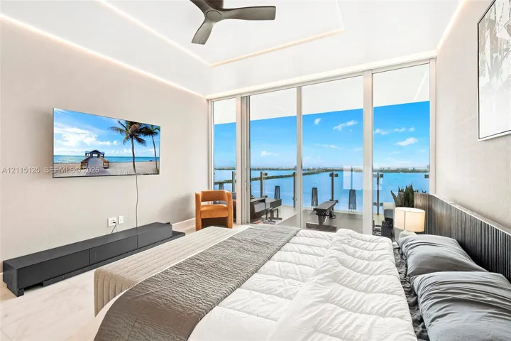 Floor-to-ceiling sliding glass doors, private balcony with glass railing, recessed tray ceiling with integrated cove lighting, matte black ceiling fan, and large-format tile flooring