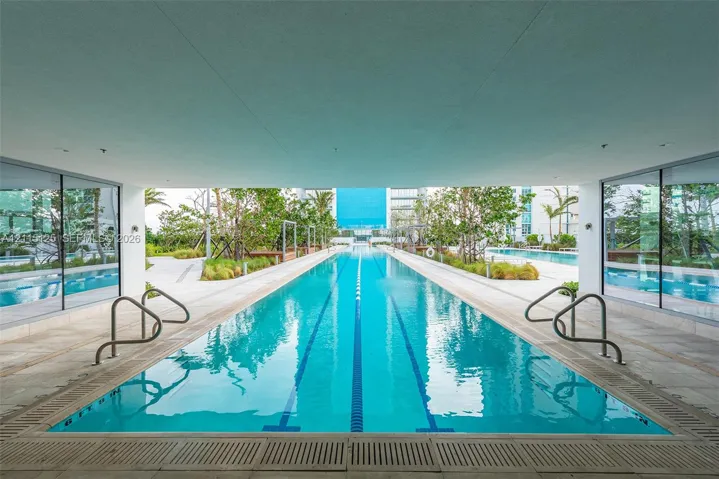 Lap pool featuring a covered perimeter, floor-to-ceiling glass wall enclosures, integrated lane markers, and stone-finished pool decking