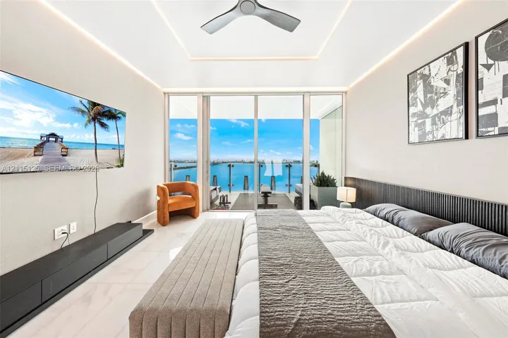 Floor-to-ceiling glass sliders, tray ceiling with recessed perimeter lighting, modern ceiling fan, dark slatted wood headboard, and polished stone flooring