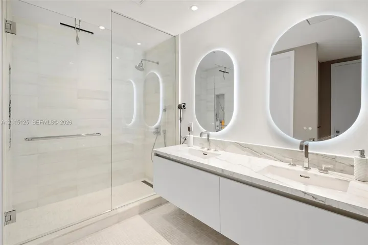 Floating dual vanity with stone countertop, integrated sinks, chrome fixtures, backlit oval mirrors, and a glass-enclosed shower featuring tile surrounds