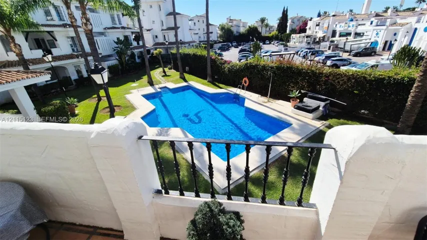 Outdoor space featuring a swimming pool with stone coping, manicured lawn, perimeter privacy hedging, and a white stucco masonry wall with decorative black metal railing