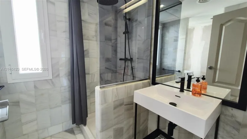 Bathroom featuring natural stone wall and floor tiling, walk-in shower with a glass panel, rainfall shower head, and a white rectangular vessel sink with matte black hardware