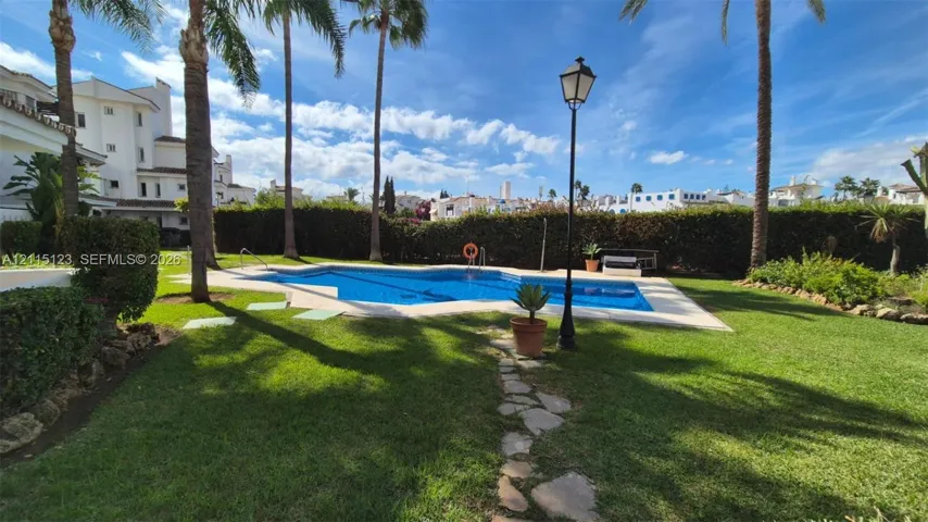 In-ground swimming pool with stone-style decking, bordered by a manicured lawn, established palm trees, and privacy-providing hedge fencing