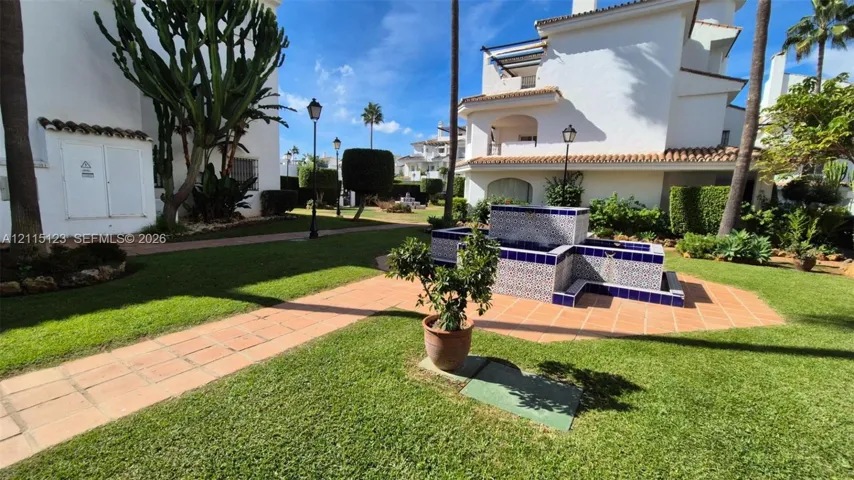 Manicured communal grounds featuring a tiered decorative fountain with patterned tile inlay, terracotta-style paving, mature palm trees, and structural landscape lighting