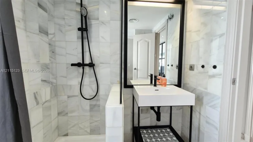 Shower stall with full-height stone wall tiling, matte black wall-mounted shower system, metal frame console vanity with white rectangular basin, and matching matte black faucet and mirror frame