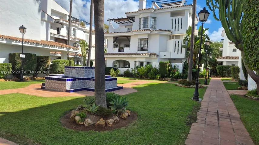 Exterior courtyard featuring a decorative tile fountain, manicured lawn, terracotta-paved pathways, mature landscaping, and multi-level buildings with integrated terraces