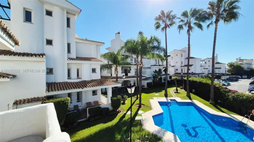 Multi-level residential exterior featuring white stucco facades, terracotta tile roofing, a communal swimming pool, landscaped lawn, and mature palm trees