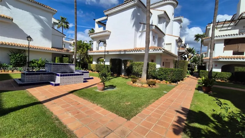 Mediterranean-style exterior with white stucco facade, terracotta roof tiles, decorative tiled fountain, paved walkways, and manicured green space