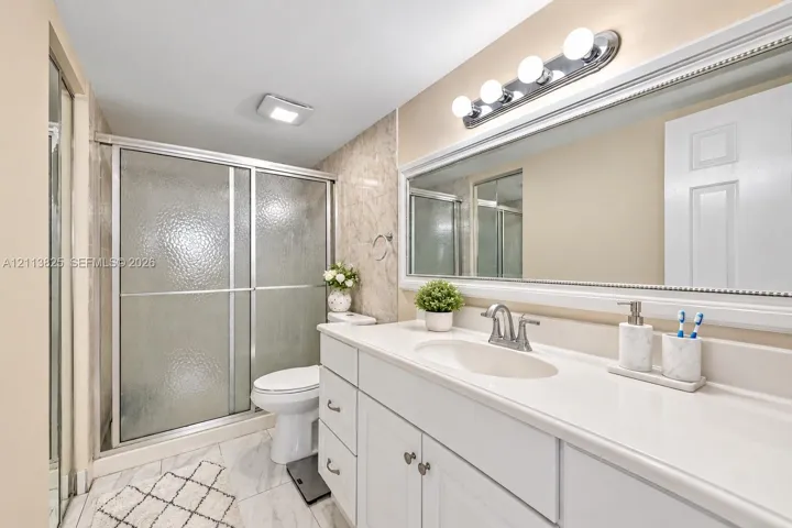 Bathroom featuring a white vanity with a solid-surface countertop, an integrated sink basin, a full-width framed mirror, a glass-enclosed shower with a sliding door, and neutral tile flooring