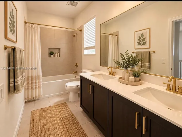 Bathroom featuring a dual-sink vanity with a solid-surface countertop, dark-finish cabinetry, brushed-gold hardware, and a tiled shower surround with an inset niche