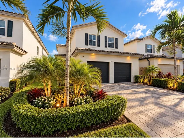 Two-story stucco exterior featuring a paver driveway, two-car garage, tile roofing, and mature palm tree landscaping
