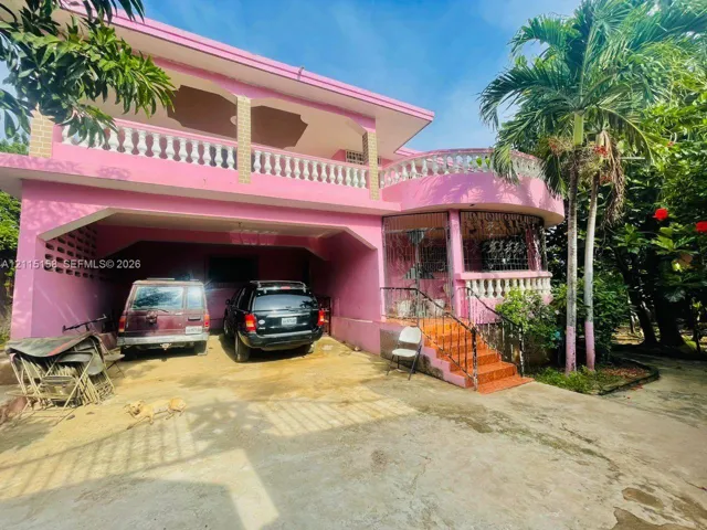 Two-story concrete residence featuring a covered carport, upper-level terrace with a balustrade, curved architectural facade, and exterior metal security grilles