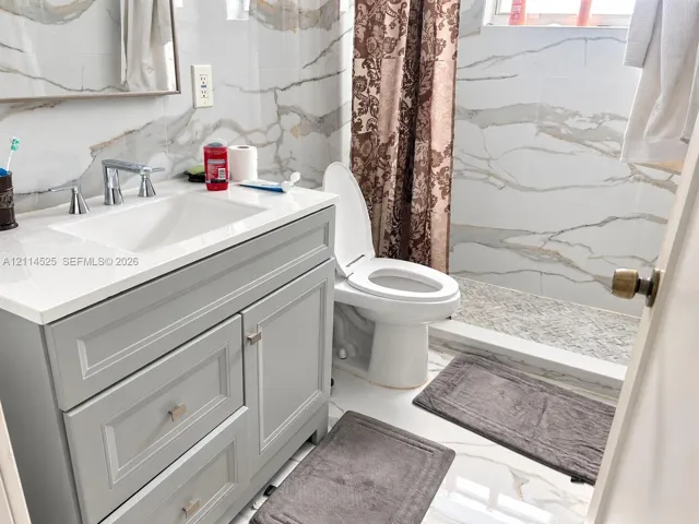 Bathroom featuring a grey shaker-style vanity with a solid surface countertop, an undermount sink with a polished chrome faucet, and large-format wall tiling with a vein pattern