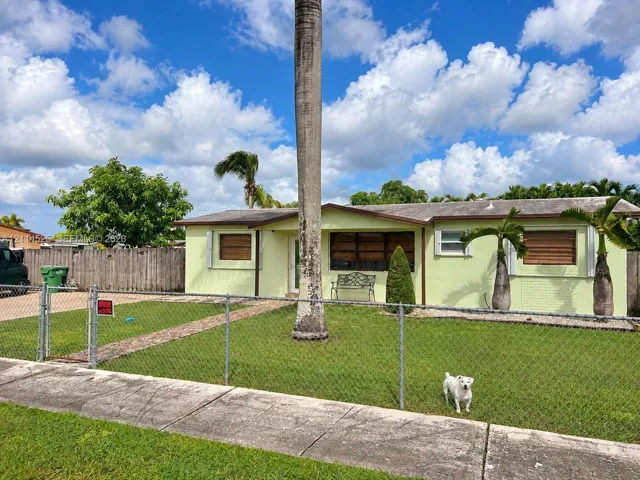 Single-story exterior featuring painted masonry siding, a gabled roofline, metal storm shutters, and a chain-link fenced front yard with mature landscaping