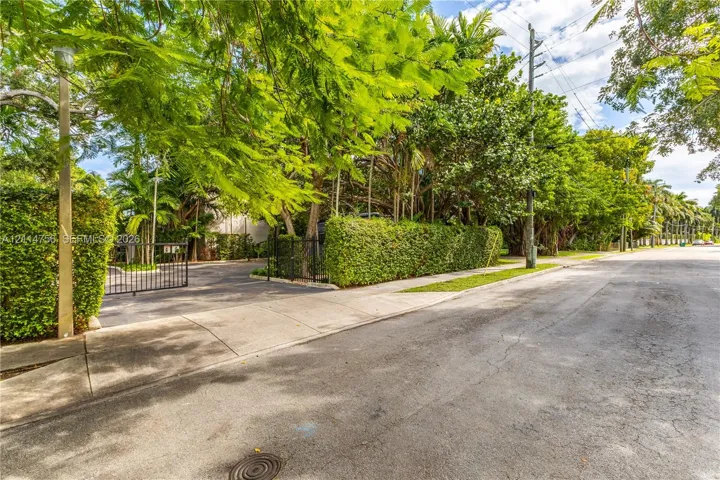 Exterior setting featuring a gated asphalt entrance, mature perimeter landscaping, concrete sidewalk, and established canopy trees