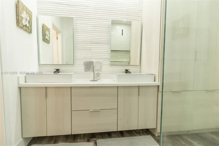 Double vanity featuring rectangular vessel sinks, light wood-tone cabinetry, a textured tiled wall accent, and frameless glass shower enclosure