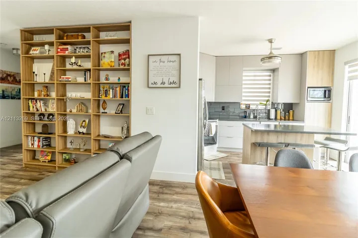 Custom built-in floor-to-ceiling shelving unit, wood-finish flooring, kitchen island with countertop seating, light grey cabinetry, and horizontal window shades