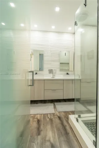 Dual-sink vanity with light wood-finish cabinetry, quartz countertops, textured tile backsplash, wood-finish flooring, and a glass-enclosed shower