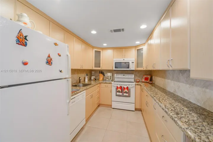 Galley-style kitchen featuring light wood cabinetry, stone-patterned countertops, a stone tile backsplash, recessed lighting, and large-format tile flooring