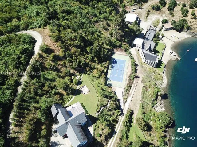 Aerial perspective of expansive coastal grounds featuring a private tennis court, waterfront access, mature woodland, and open lawn areas