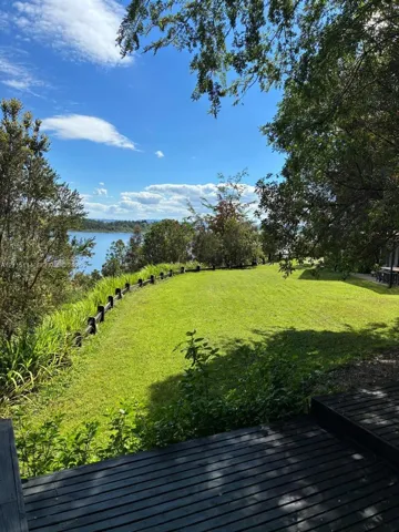Expansive lawn area featuring a dark-toned wood-plank deck, perimeter timber post-and-rail fencing, mature trees, and water scenery