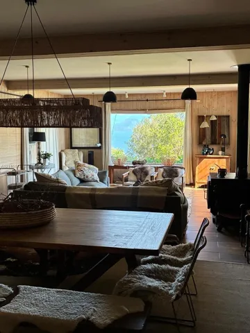 Wood-paneled interior featuring exposed structural beams, a wood-burning stove, large picture window with water scenery, and suspended pendant lighting