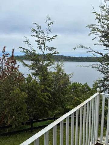 Waterfront parcel featuring a white railing, black split-rail fencing, manicured grass, and mature trees